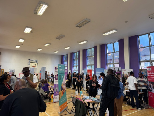 A room of people, with purple curtains. People standing at stalls and chatting.